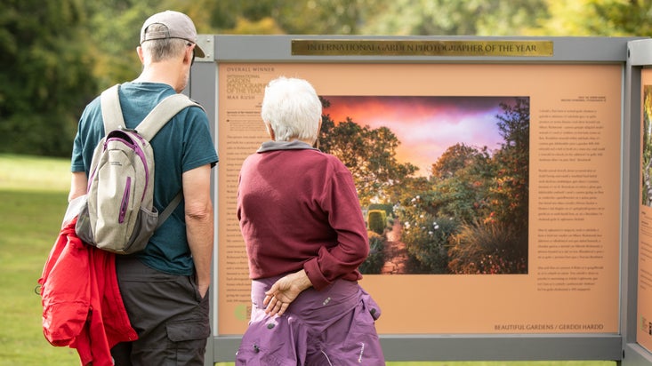 Two people looking at an interpretation board with a large photo of a garden at sunset on it as part of the International Garden Photographer of the Year exhibition at Bodnant Garden